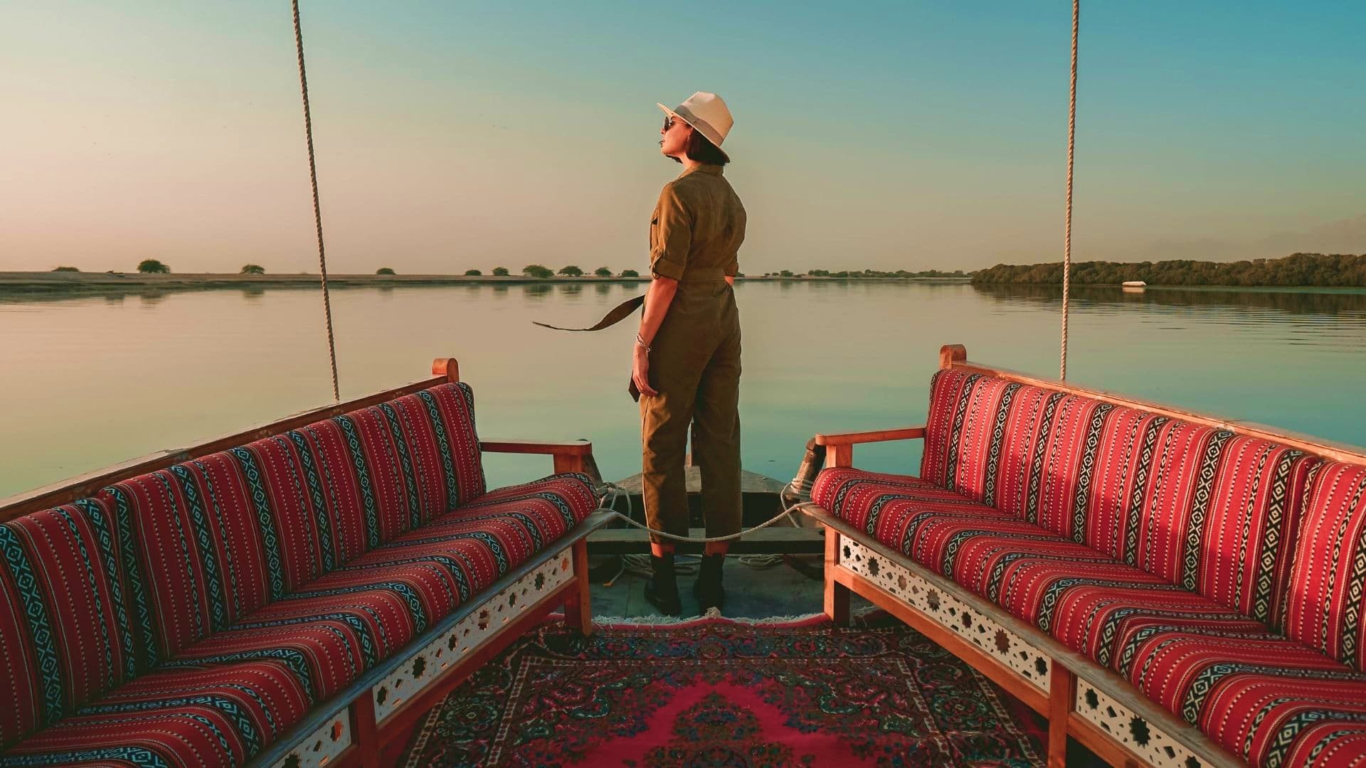 Man in safari outfit standing on wooden boat with red patterned cushions on calm river at sunrise